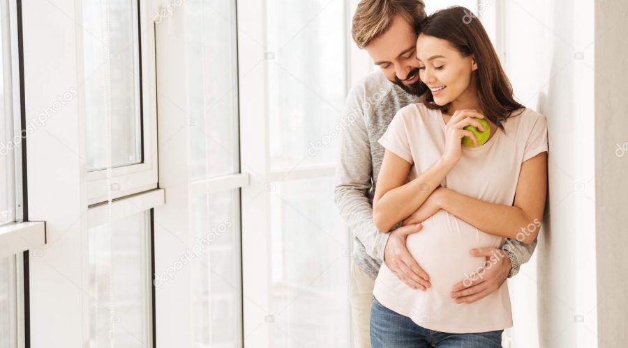 Smiling pregnant young couple hugging while standing together at a window